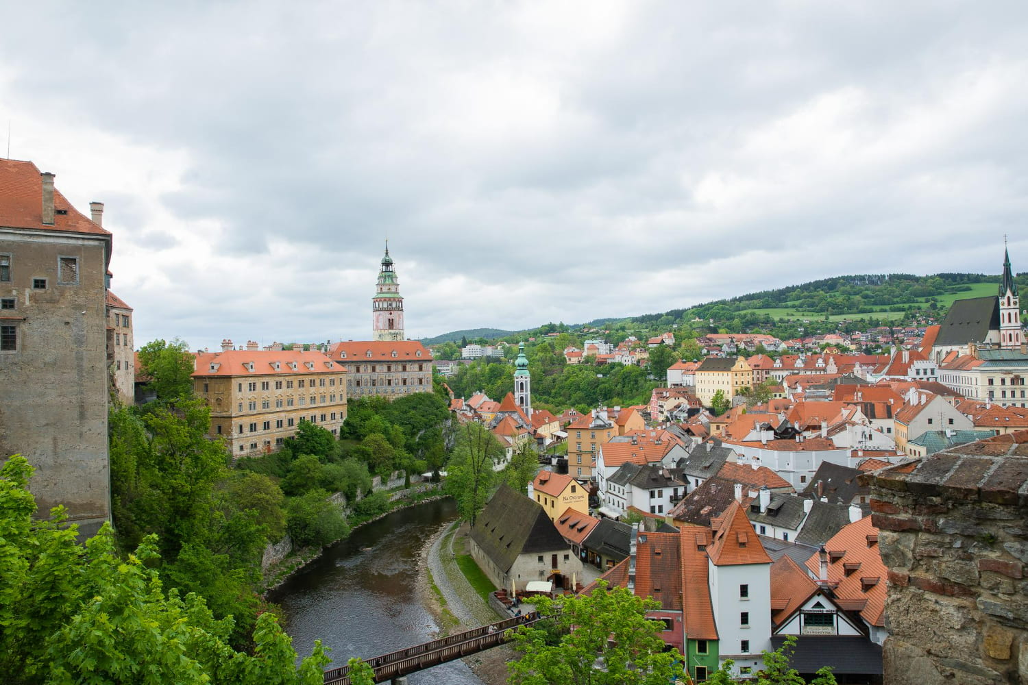 A spectacular panoramic view captures a picturesque medieval European town nestled along a winding river, surrounded by rolling green hills.