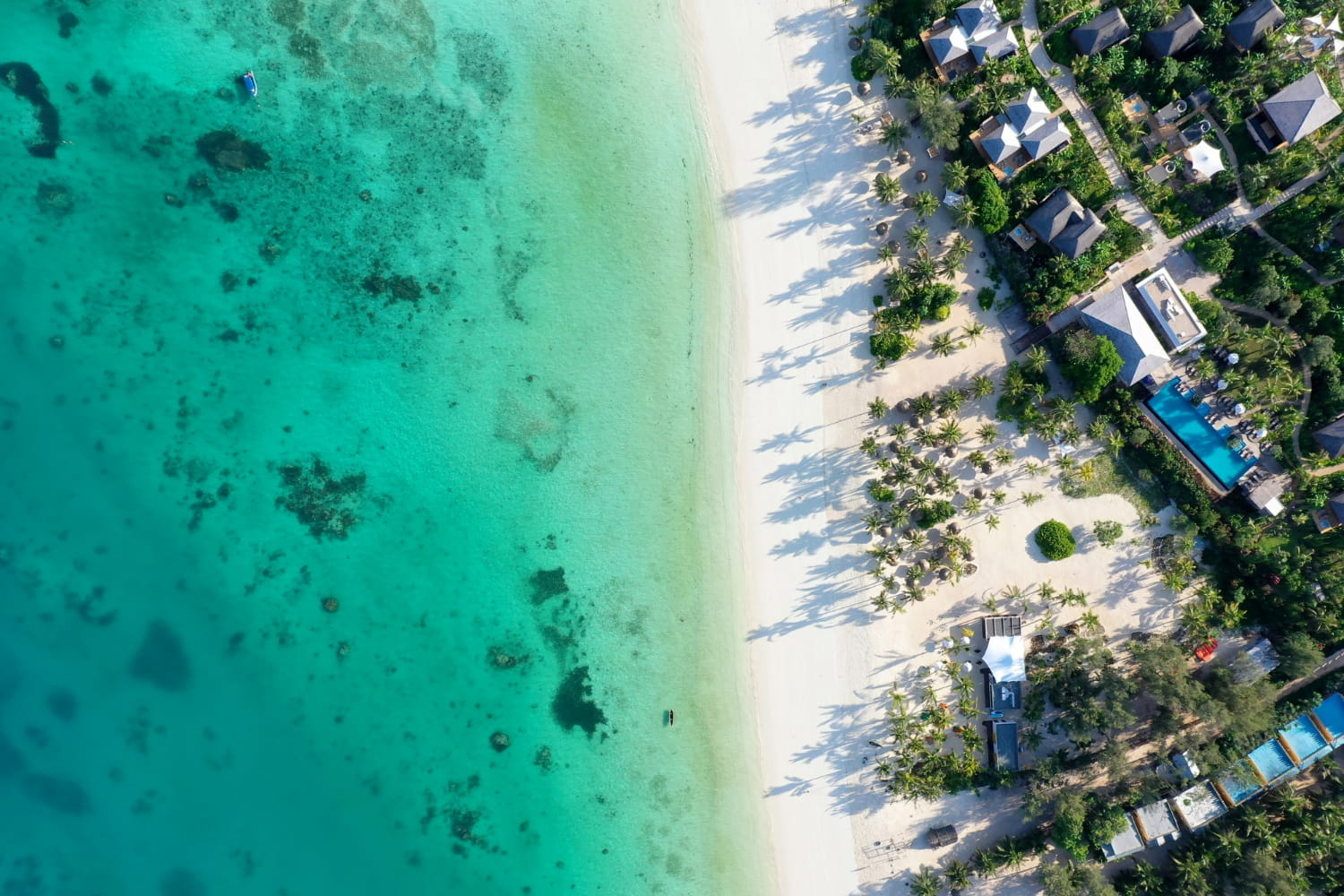 This breathtaking aerial photograph captures a luxurious tropical resort nestled along a pristine white sand beach, framed by stunningly clear turquoise waters.