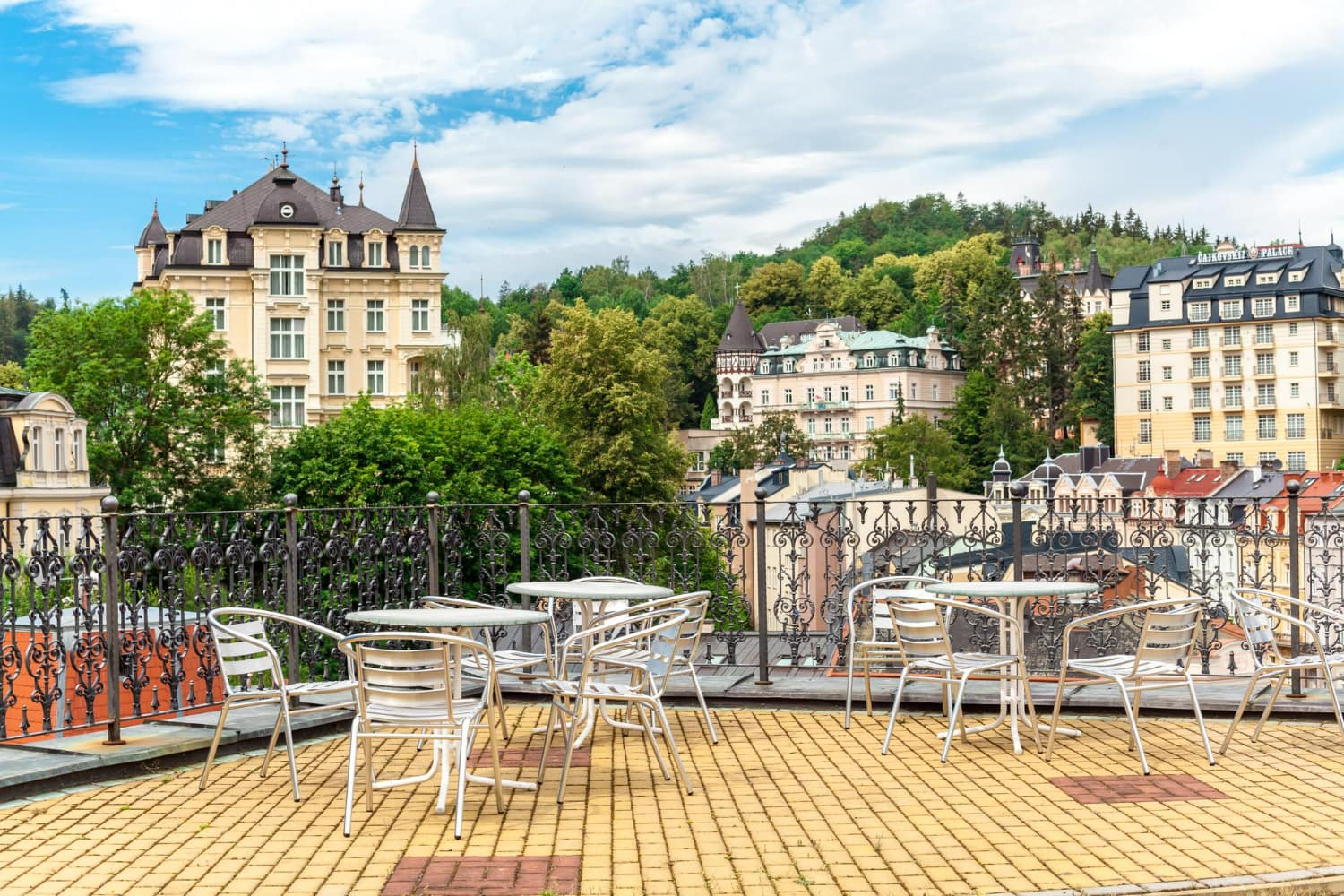 A sophisticated outdoor terrace overlooking a picturesque Central European spa town features comfortable seating arrangements with white tables and chairs arranged for leisurely relaxation.