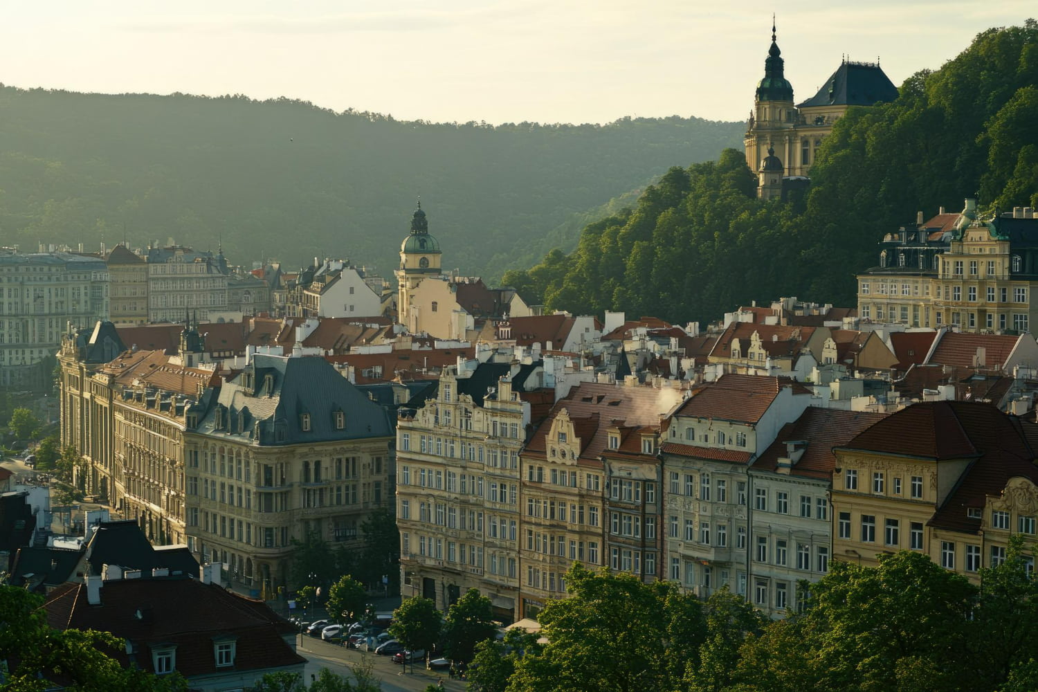 A breathtaking aerial view captures a picturesque Central European town nestled within a lush, forested valley surrounded by rolling green hills.